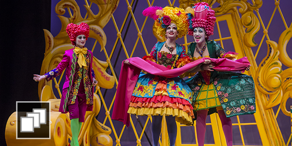 Three opera singers wearing brightly colored costumes and wigs performing on stage.
