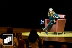 Tina Fey sitting on stage while holding a microphone and laughing.