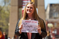 A woman in a black jacket holding a sign in front of Temple’s bell tower.